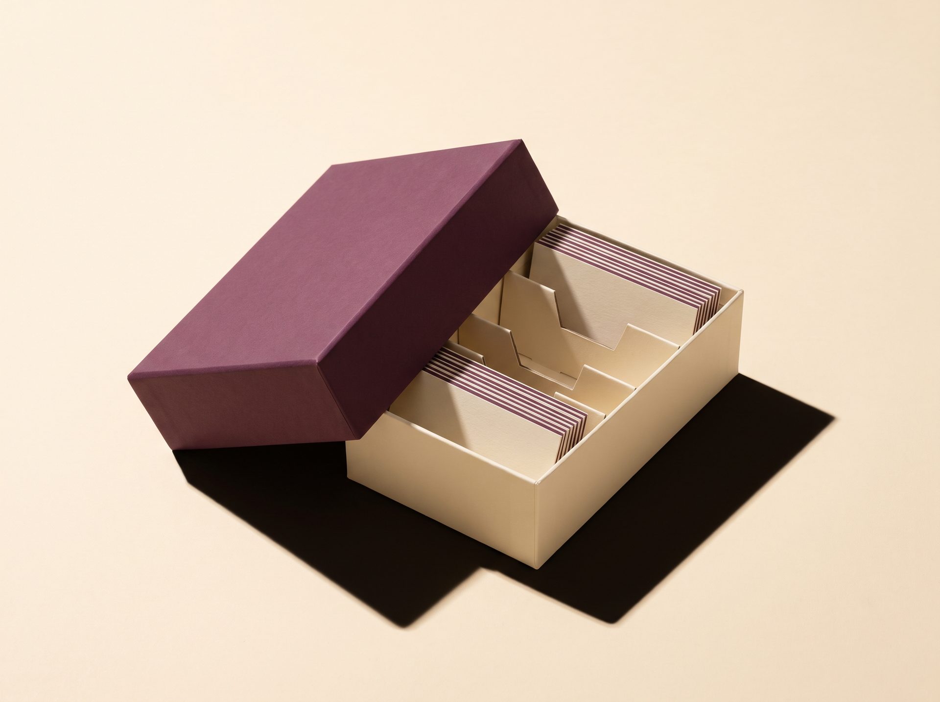 A single boxed kit with a plum lid, tilted on warm cream paper, casting a hard shadow