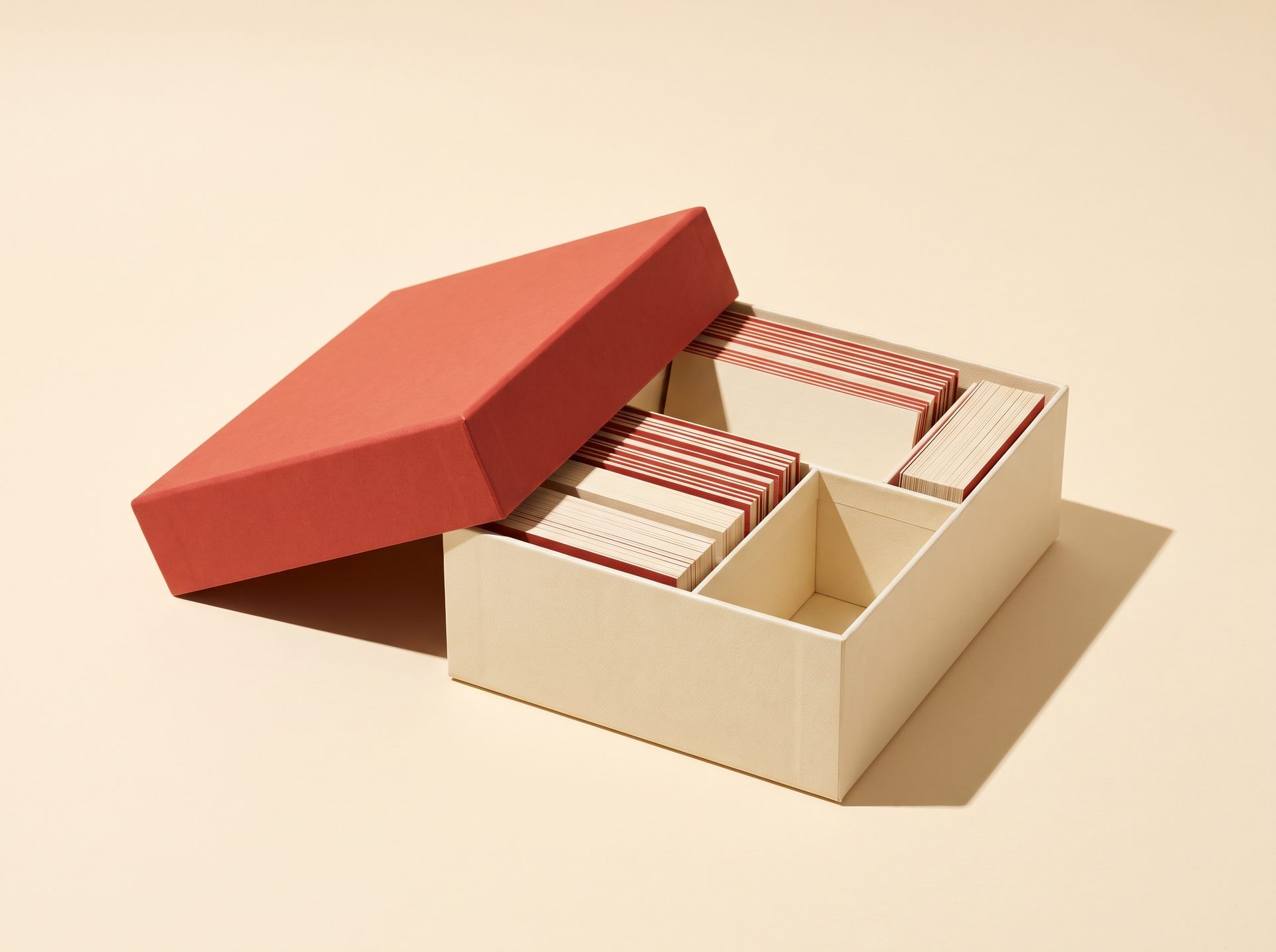 A boxed kit with a warm red lid tilted open on cream paper, casting a hard shadow