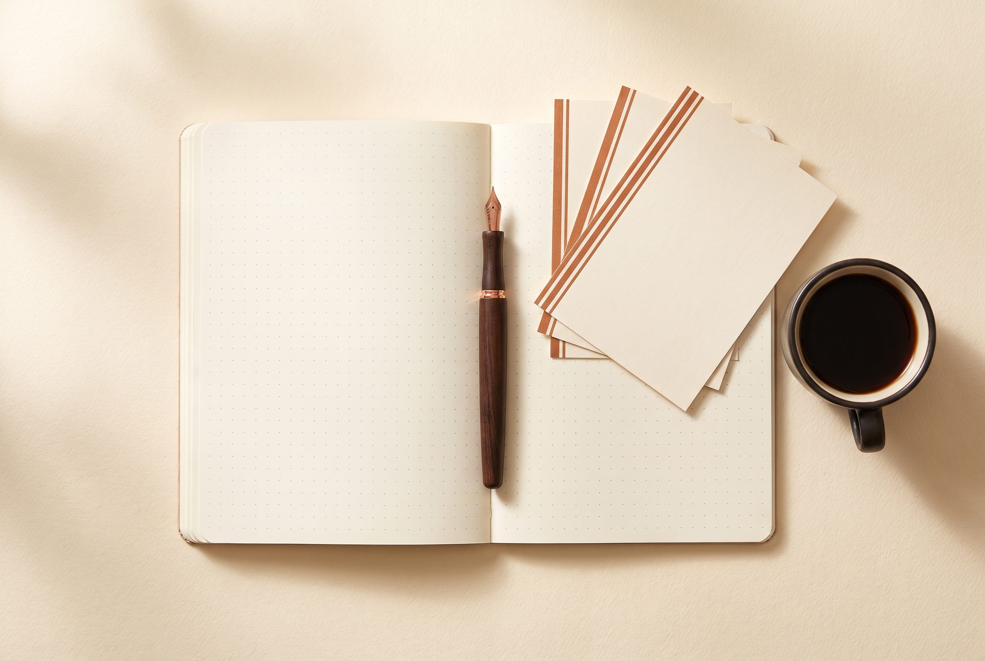 An open blank dot-grid notebook on warm cream paper, with a pen, a stack of copper-edged cards and a cup of coffee beside it